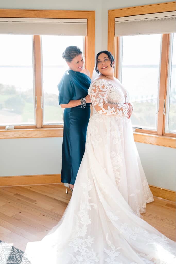 Plus Size Bride wearing a long-sleeve lace A-line wedding dress being helped by her mother during getting-ready photos, natural light coming through large windows. Bridal gown from Blue Violet Bridal Prom & Tux a size-inclusive shop serving Eastern CT and Southern RI.