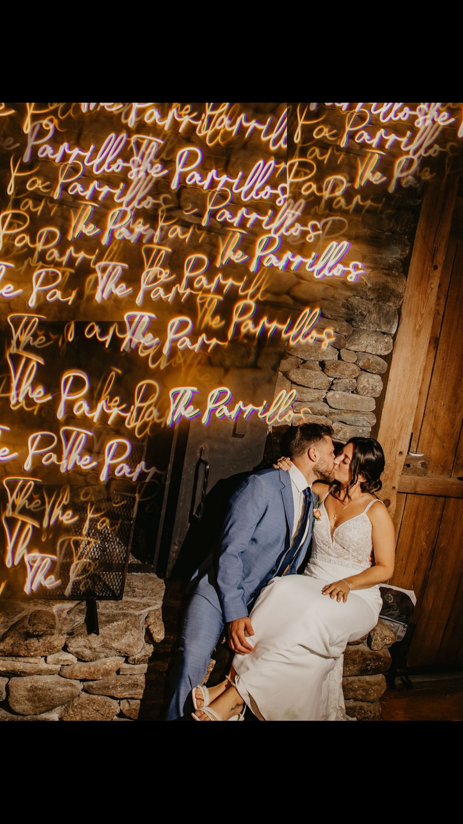 Bride and groom sharing a kiss at their wedding reception in front of a neon “The Parrillos” sign, bride wearing a modern crepe wedding gown with lace detailing. Gown from Blue Violet Bridal Prom & Tux.